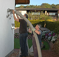 Zwei Männer, einer steht auf einer Leiter, der Andere hält einen langen dicken Schlauch, der an die weit oben an der Hauswand  gelegene Einblaskupplung angeschlossen werden soll 